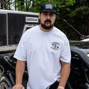 A man wearing a white Hoonigan LOW COMPRESSION short sleeve t-shirt stands next to a car, showcasing automotive lifestyle apparel.
