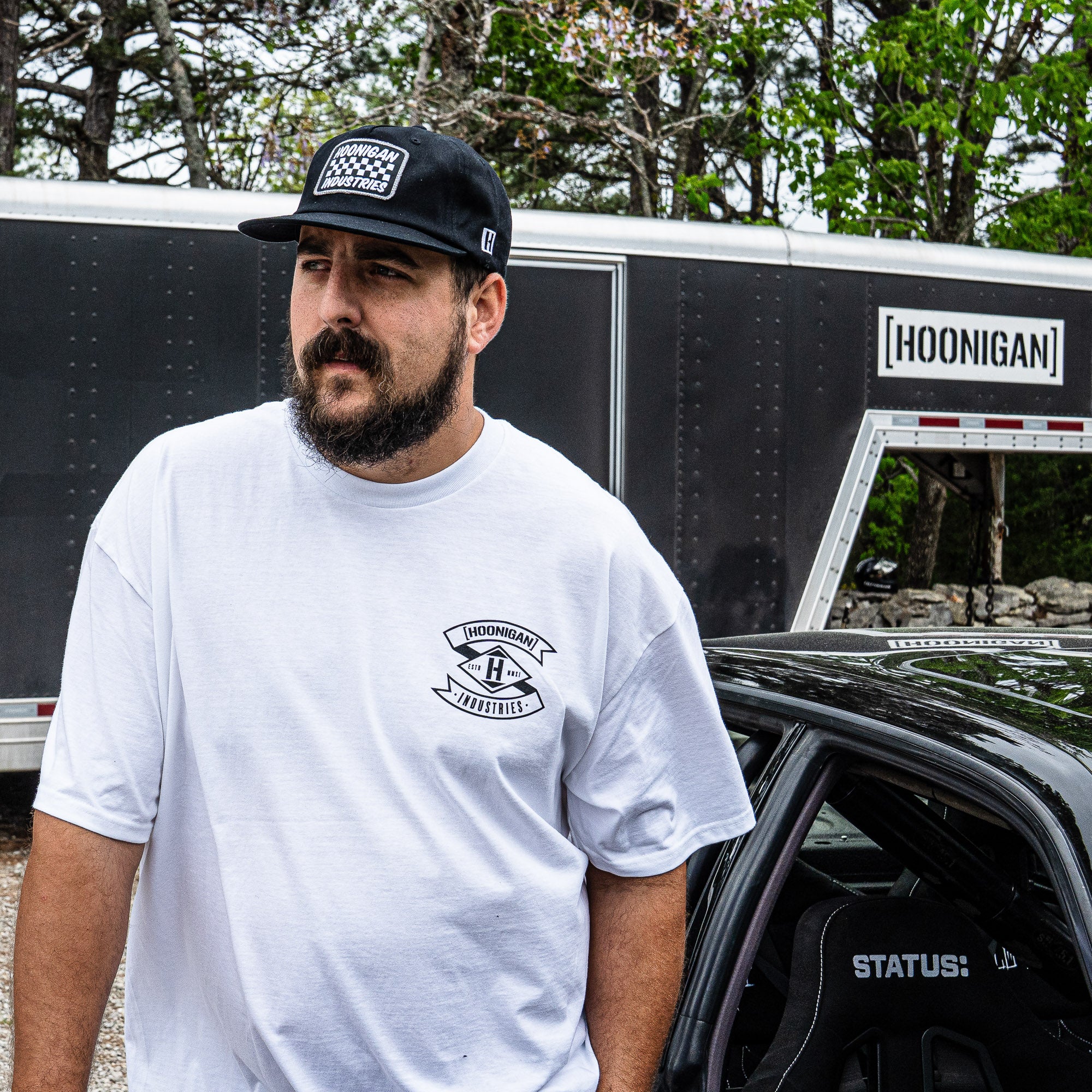 Man wearing a Hoonigan LOW COMPRESSION short sleeve t-shirt stands next to a car, showcasing automotive lifestyle apparel.