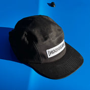 Black unstructured strapback hat featuring a white Hoonigan logo patch, resting on a blue surface.