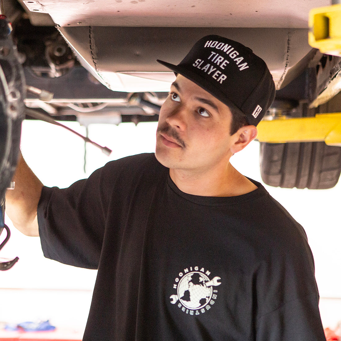 Hoonigan TIRE SLAYER Snapback Hat featuring a black design with bold white text, set against a casual outdoor background.