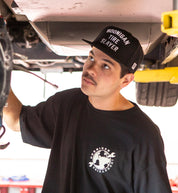 Hoonigan TIRE SLAYER Snapback Hat featuring a black design with bold white text, set against a casual outdoor background.