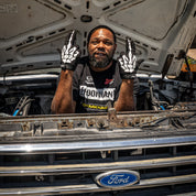 A mechanic wearing Hoonigan FIND OUT Gloves poses with a playful gesture under the hood of a Ford truck.