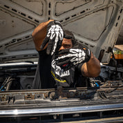 A person wearing Hoonigan FIND OUT Gloves poses with hands shaped like a skeleton while working on a car engine.