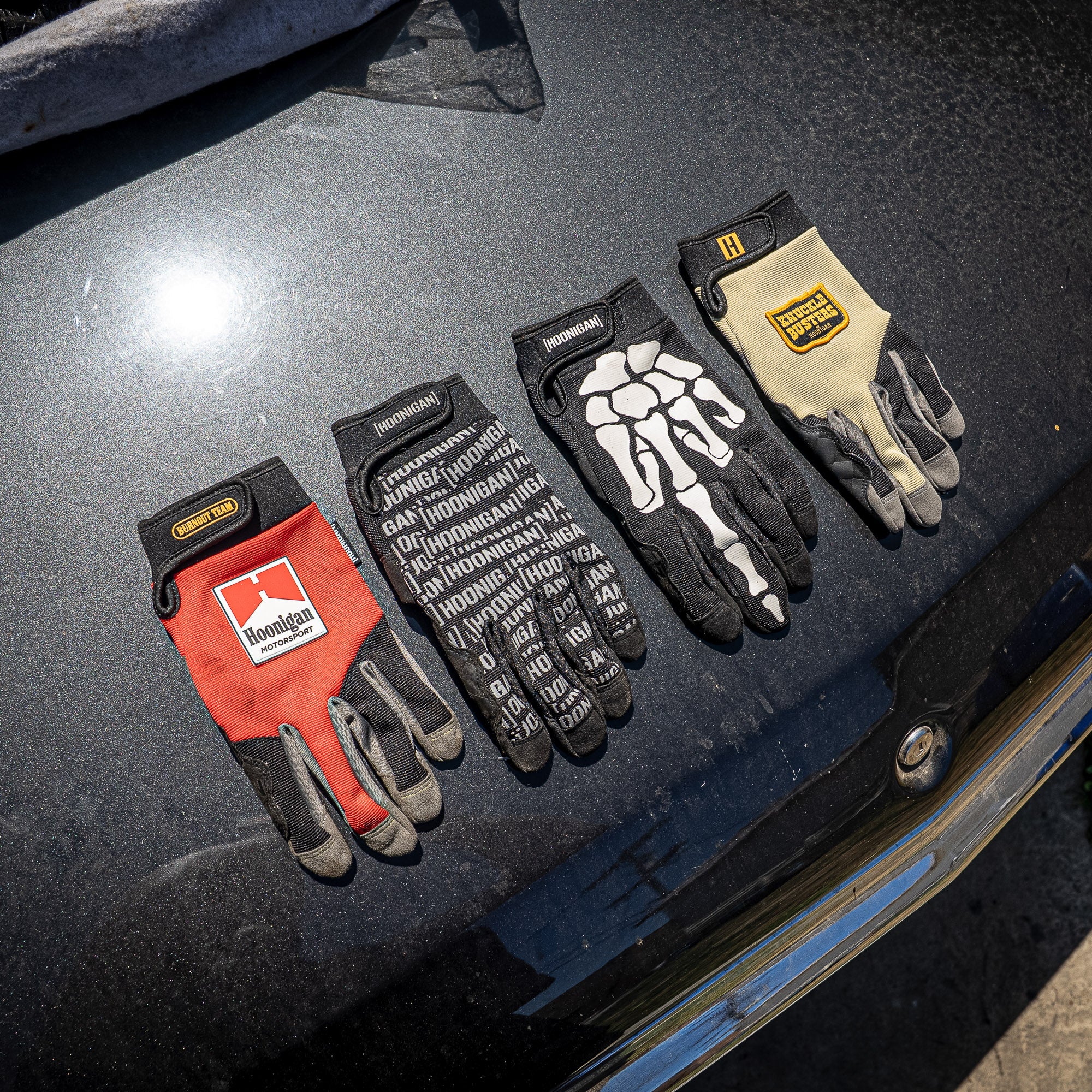 Hoonigan Surgeon General gloves displayed on a car hood, featuring various designs and colors for motorsport enthusiasts.