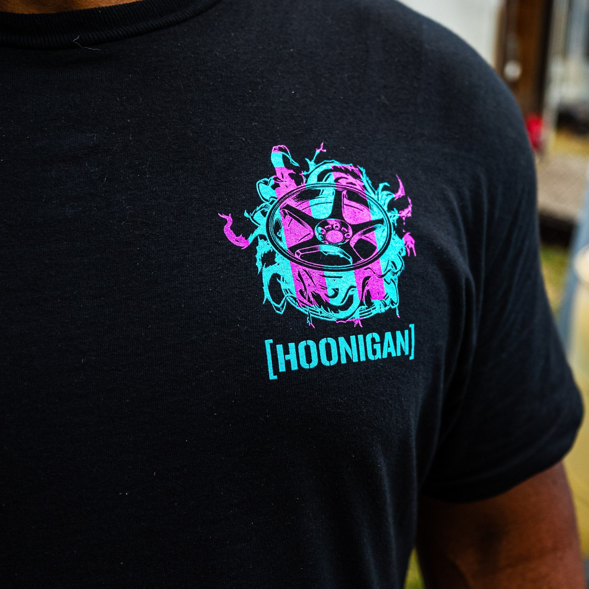 Front of the black Hoonigan TREADWEAR tee with a pink and teal wheel graphic and [HOONIGAN] logo on the chest.