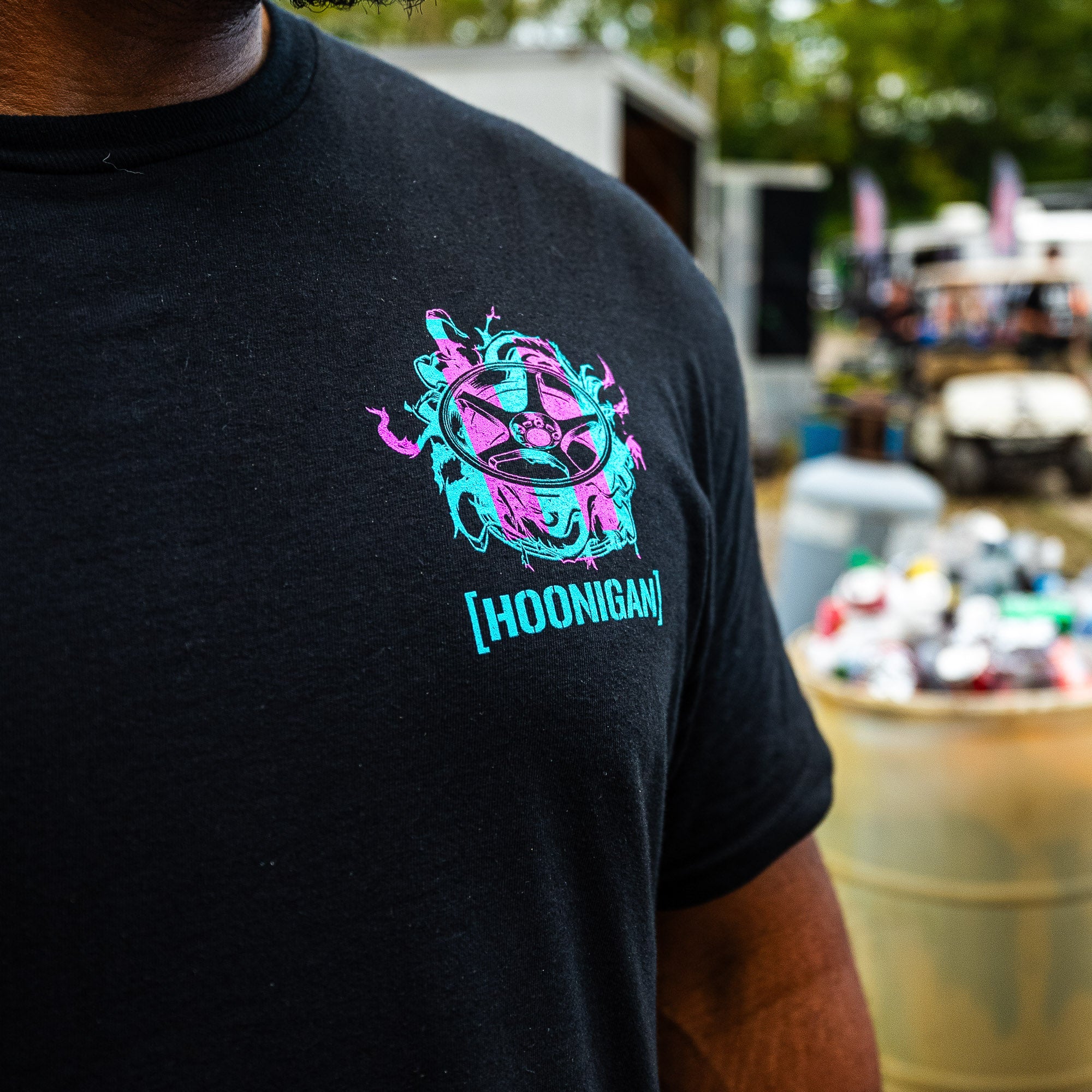 Black Hoonigan TREADWEAR tee worn at a car event, pink and teal wheel graphic on the left chest.