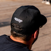 American Racing Logo Snapback Hat displayed on a flat surface, featuring a black cap with a prominent white embroidered logo on the front.