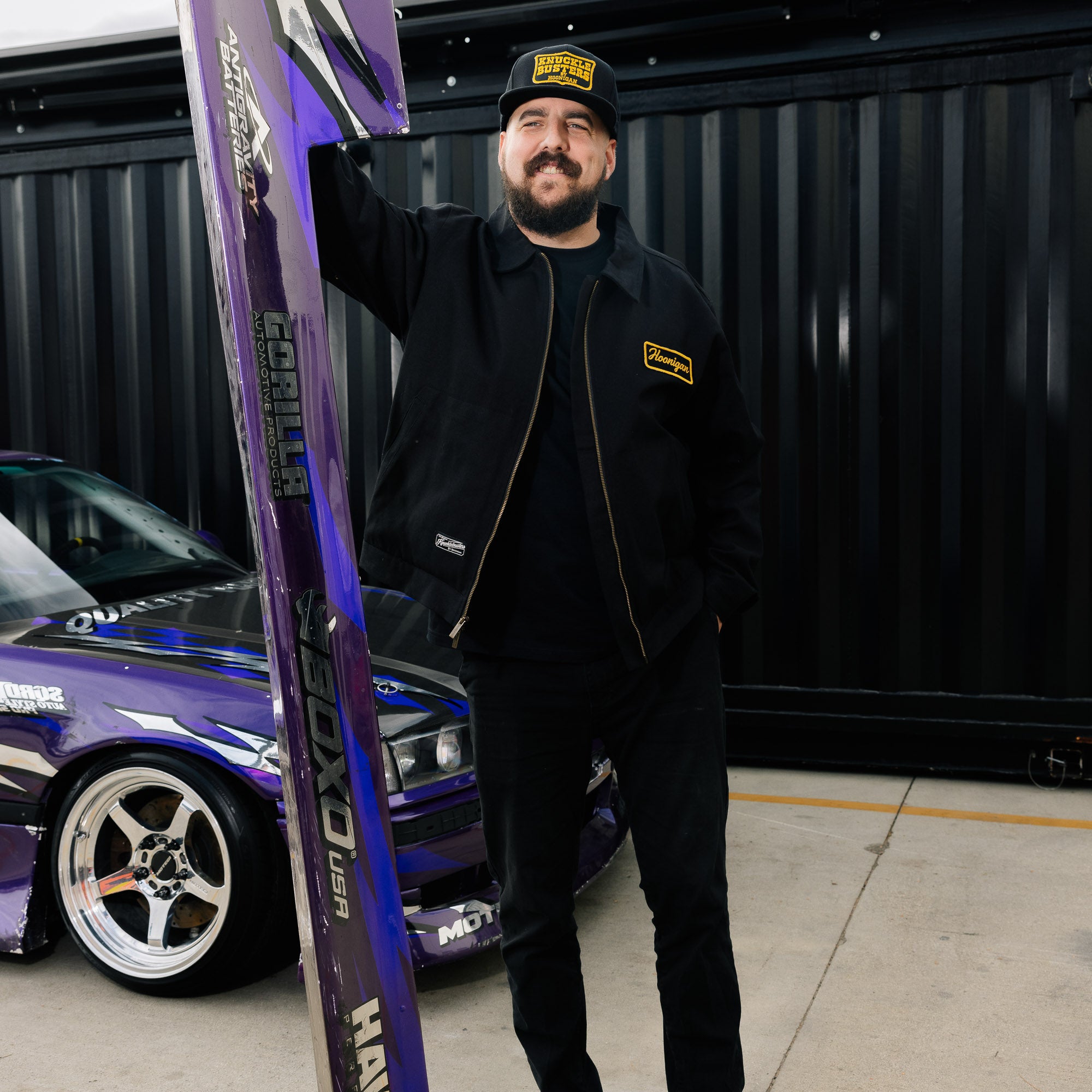 Black Hoonigan KNUCKLEBUSTERS jacket featuring a bold logo on the back and contrasting sleeves, designed for streetwear enthusiasts.