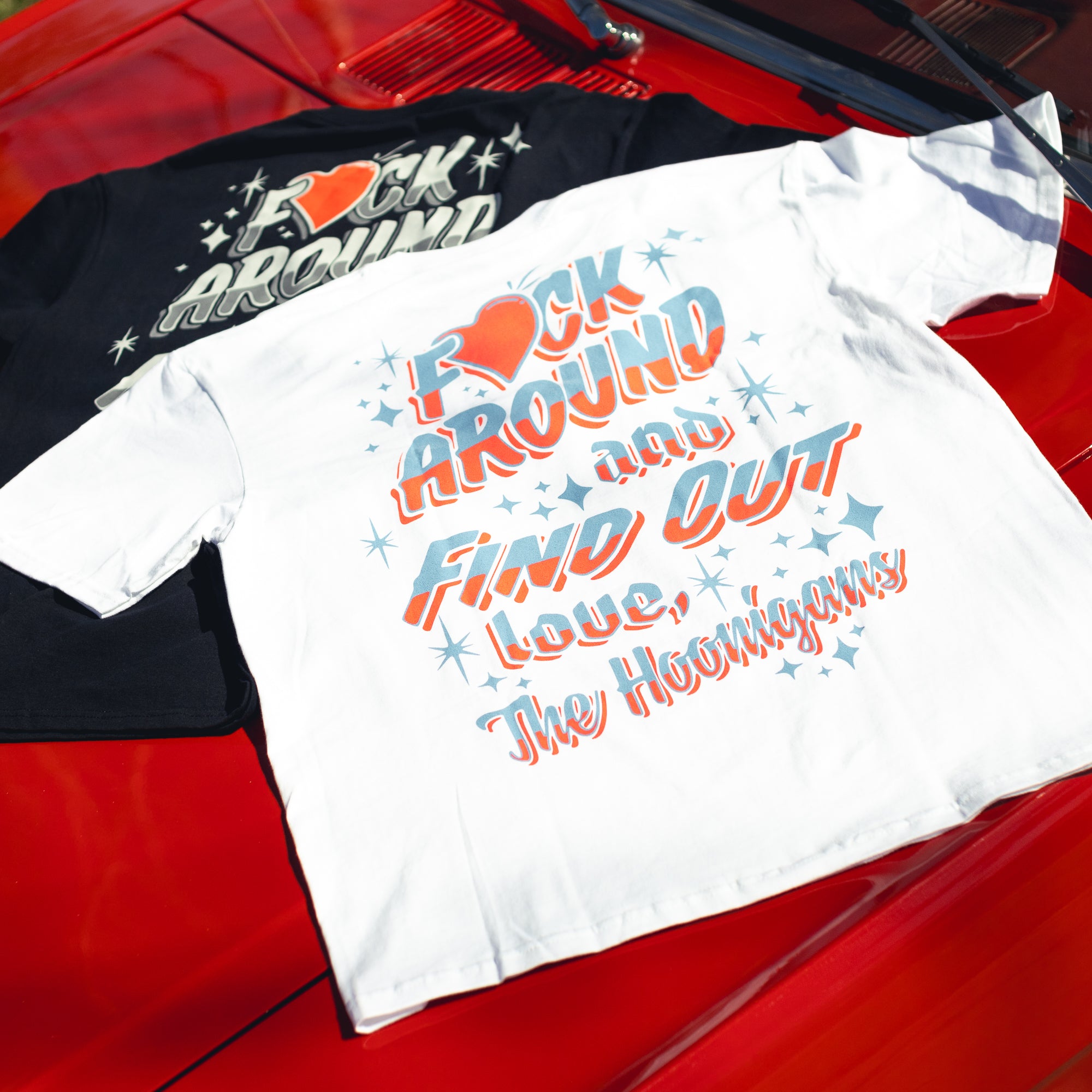 White and black Hoonigan FIND OUT V2 short sleeve tees with colorful, retro-style text and star graphics, laid on a red surface.