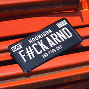 Black license plate with white text reading "FCK ARND" and "and find out," mounted on an orange surface.