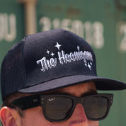 Black trucker hat with "The Hoonigans" embroidered in white, featuring stars, worn with sunglasses.