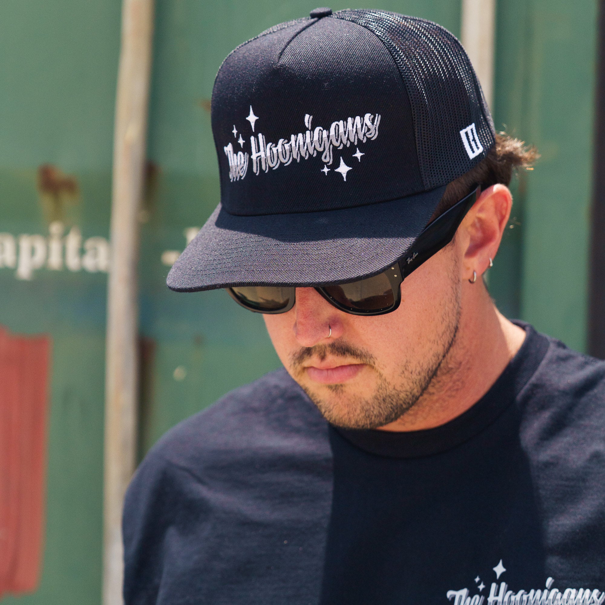 Man wearing a black snapback trucker hat with "The Hoonigans" embroidered in white, paired with sunglasses and a black T-shirt.