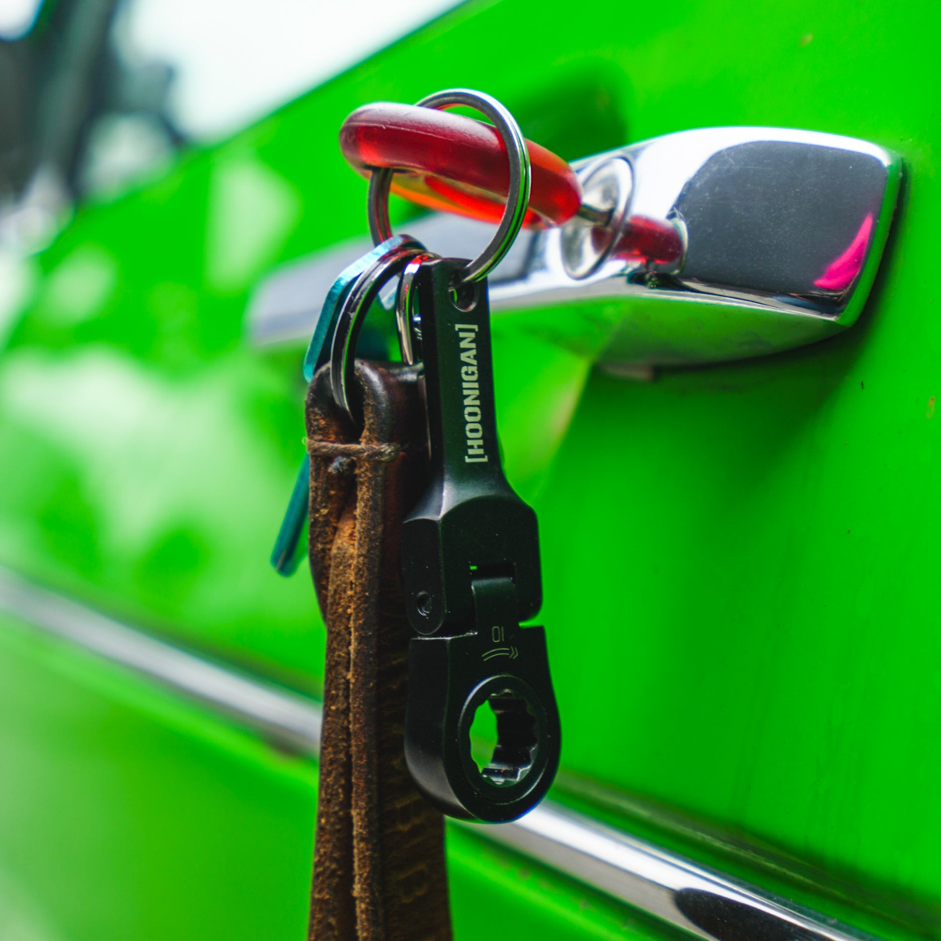 Hoonigan FOUND IT Keychain attached to a leather strap, hanging from a chrome car door handle on a vibrant green vehicle.