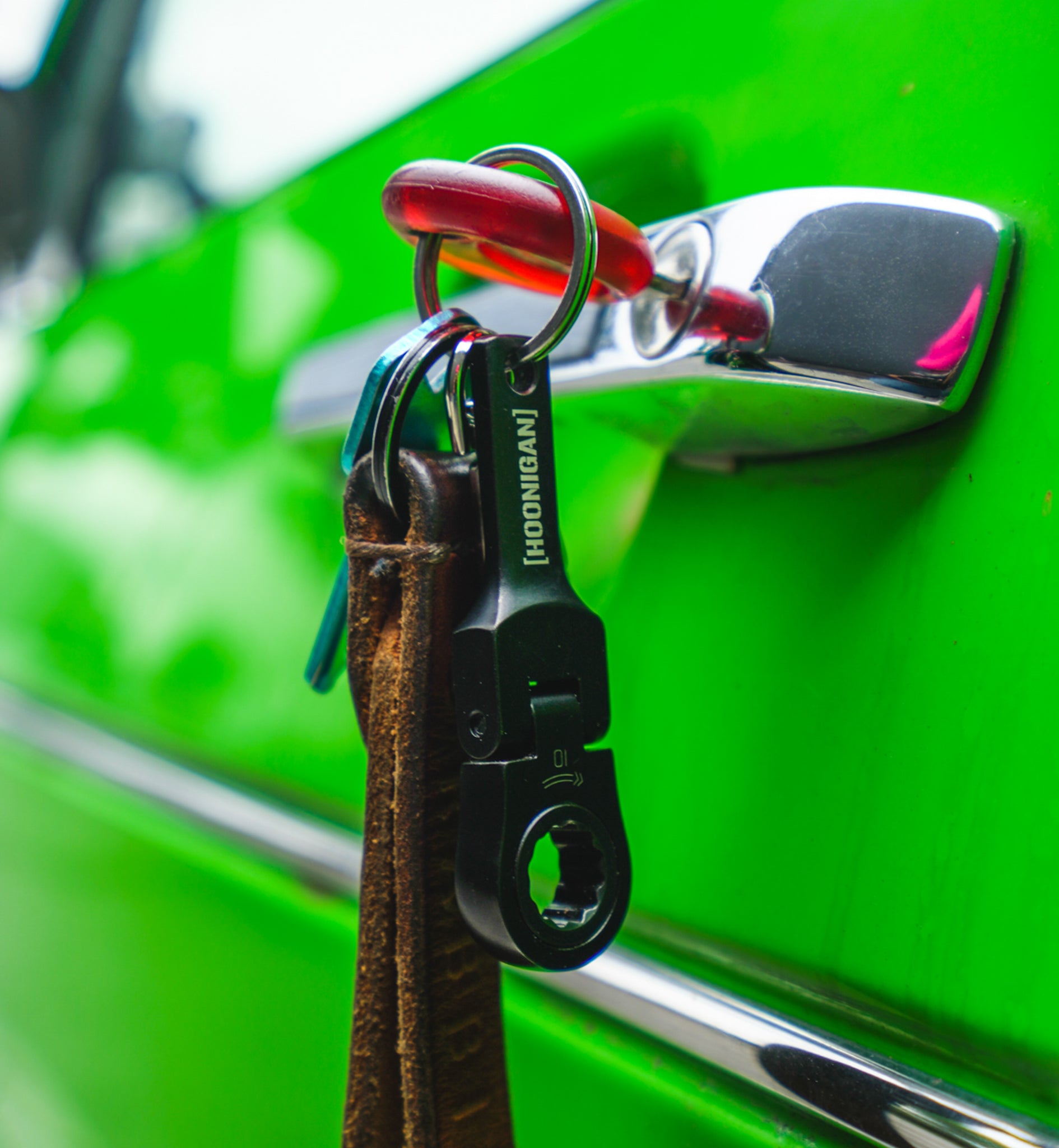 Hoonigan FOUND IT Keychain attached to a leather strap, hanging from a chrome car door handle on a vibrant green vehicle.