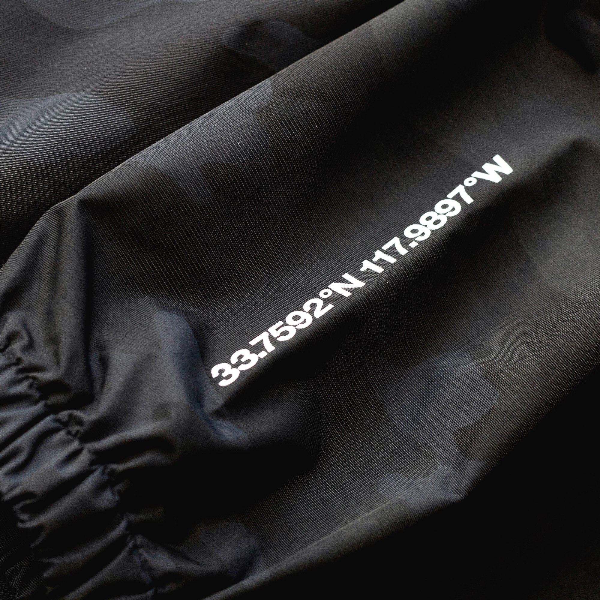 Close-up of a black Rotiform GLOBAL IMPACT half zip anorak jacket with white printed coordinates and text on the fabric.