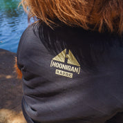 Close-up of a black Hoonigan T-shirt with a gold topographic mountain graphic and "HOONIGAN" and "HAVOC" text, worn outdoors near water.