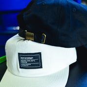 Black and white unstructured strapback hats with "Making Cool S#*% Since 09" label, featuring adjustable strap and metal buckle.