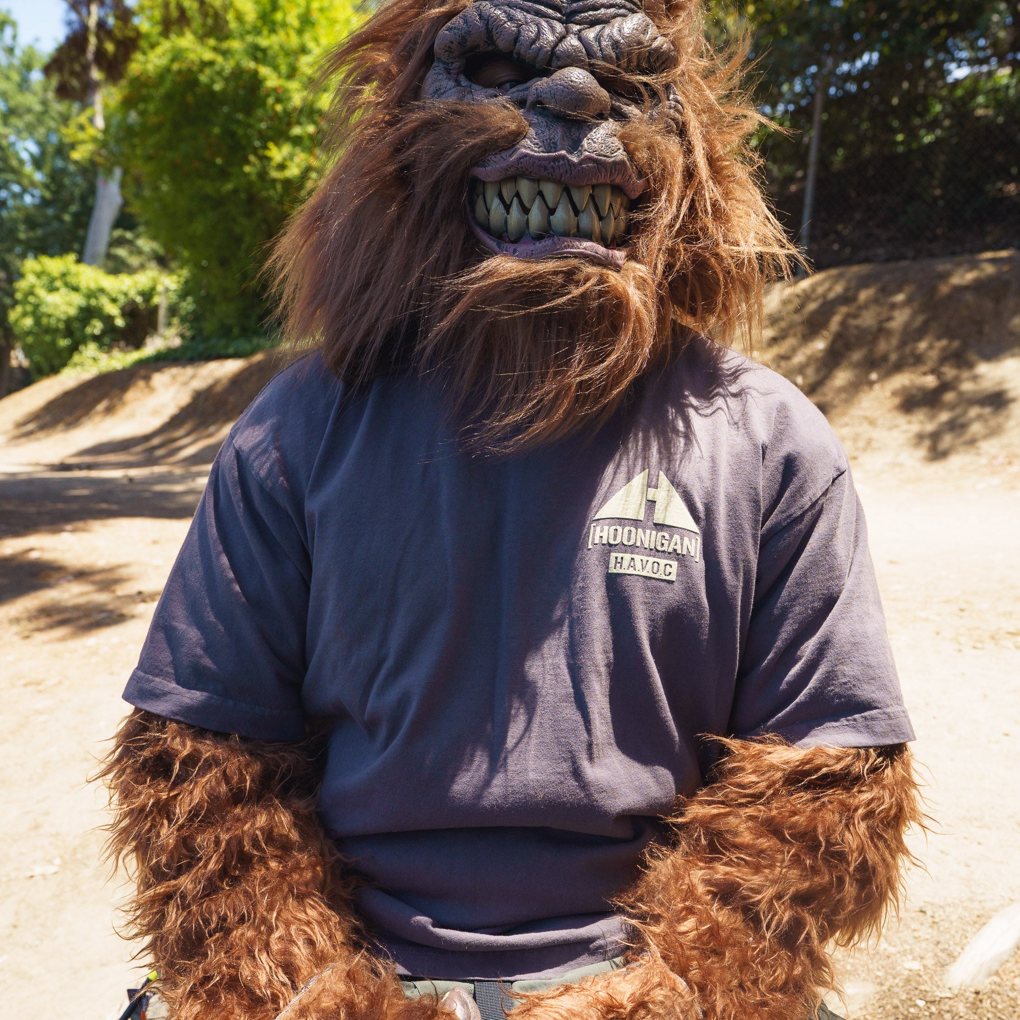 A person in a lion mask wearing a dark gray Hoonigan TOPOGRAHPHIC TERRAIN short sleeve tee outdoors on a sunny day.