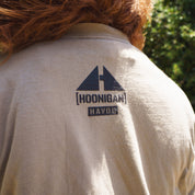 Close-up of a beige T-shirt with "Hoonigan" and "H.A.V.O.C" logos, featuring a graphic of a mountain top, worn outdoors.
