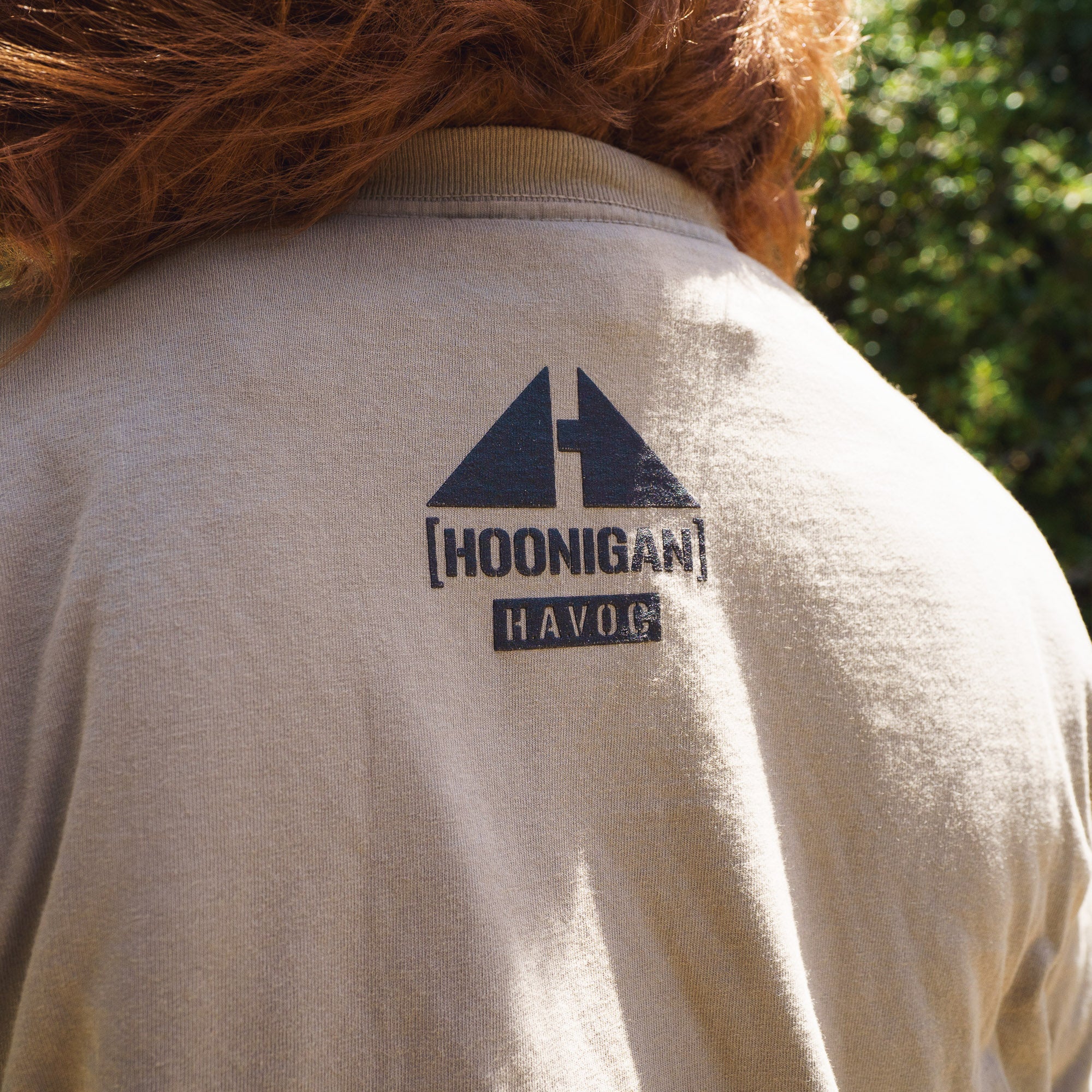 Close-up of a beige T-shirt with "Hoonigan" and "H.A.V.O.C" logos, featuring a graphic of a mountain top, worn outdoors.
