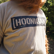 Close-up of a beige T-shirt with "Hoonigan" graphic and topographic design, worn outdoors with sunlight and greenery.