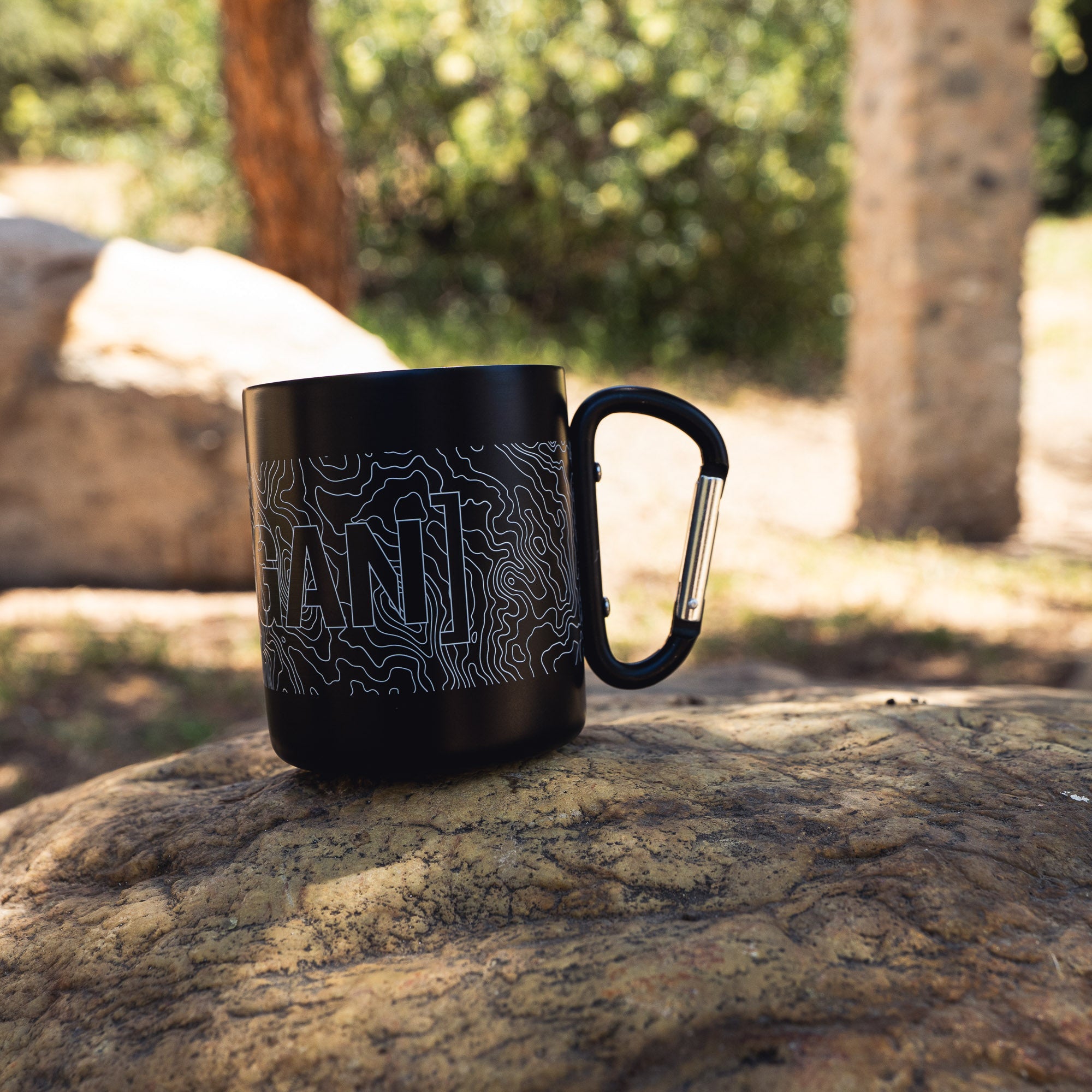 Black mug with topographic map design and the word "GAND" printed on it, featuring a carabiner handle for outdoor use.
