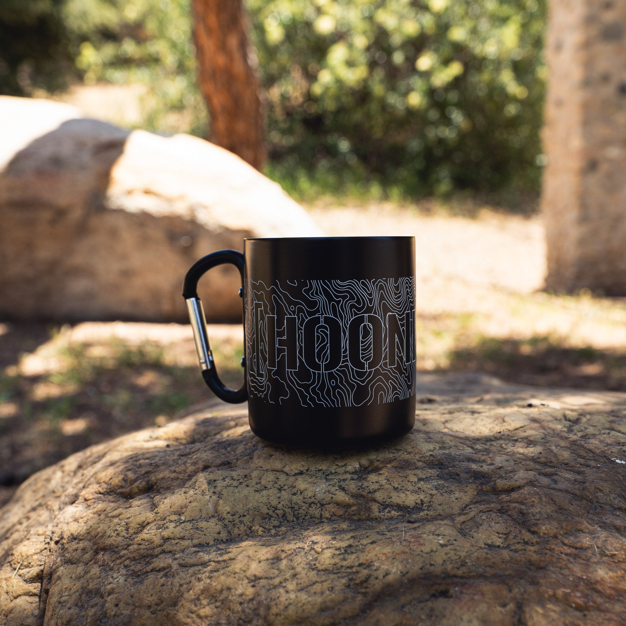 Black ceramic mug with a carabiner handle featuring topographic line design and the word "HOONIGAN" printed on it.