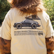 Close-up of a person wearing a beige Hoonigan SAFARI Short Sleeve Tee featuring a black vintage car with a roof tent and overland graphic design.