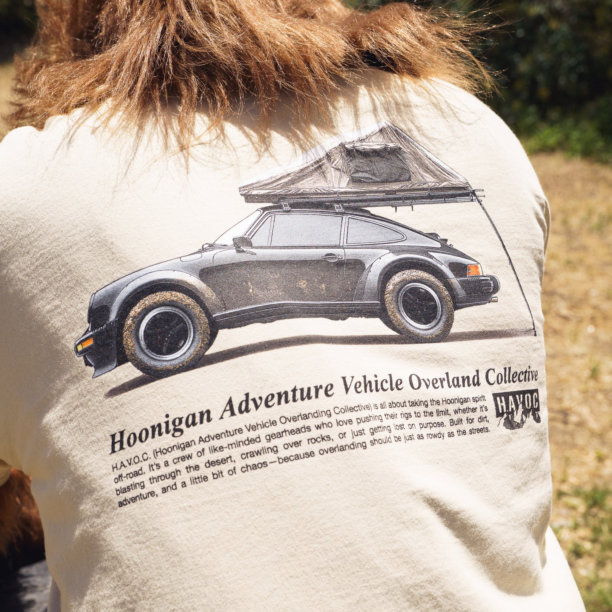Back of a beige T-shirt featuring a graphic of a vintage car with a rooftop tent, and text about the Hoonigan Adventure Vehicle Overland Collective.