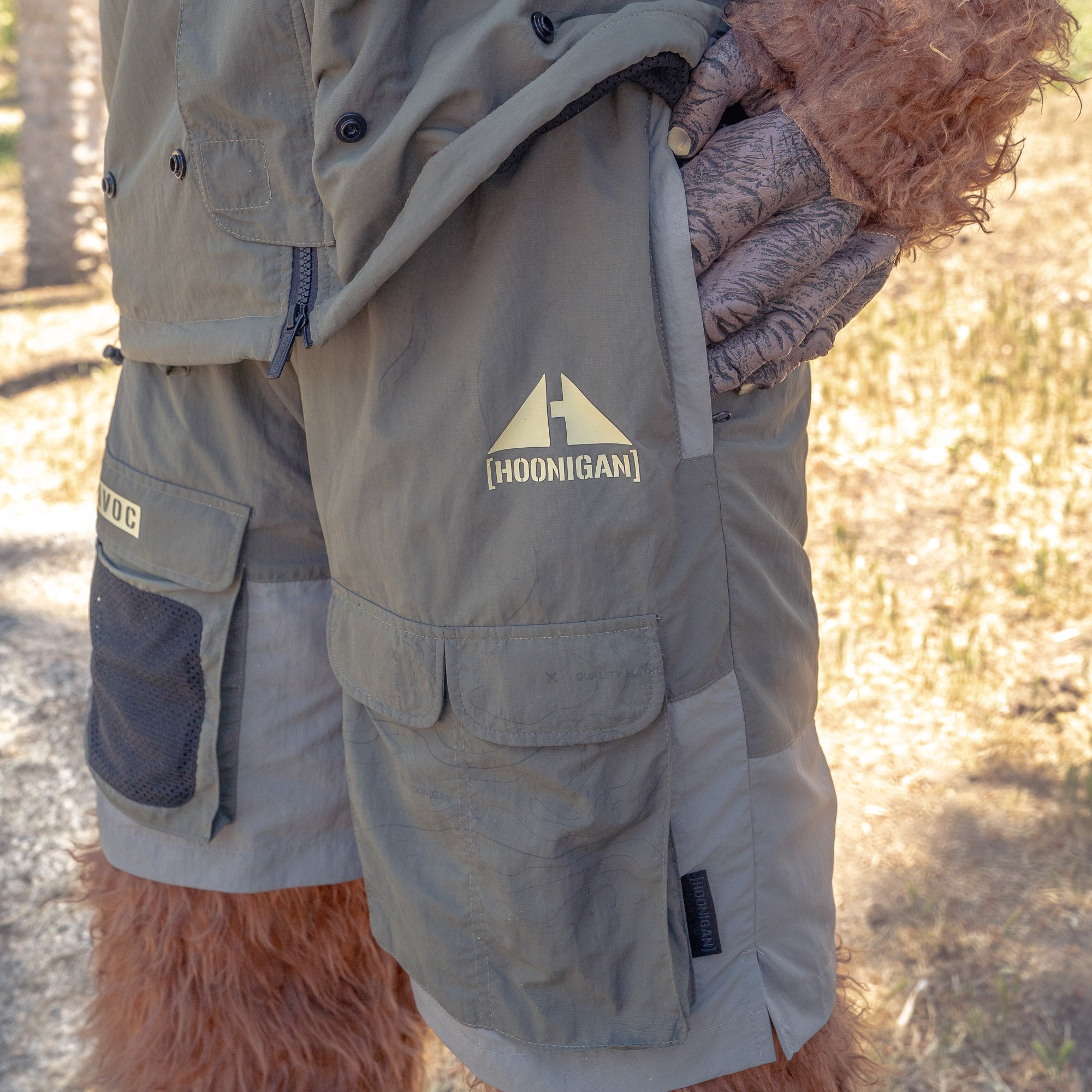 Close-up of gray Hoonigan H.A.V.O.C. rain-activated cargo shorts with multiple pockets, worn outdoors by a person with furry arms.