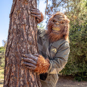 A person in a hairy monster mask and rain jacket hugs a tree outdoors on a sunny day.