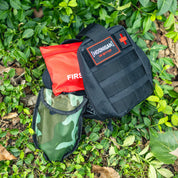 Black Hoonigan OH SH*T Safety Kit bag with red cross patch, red first aid pouch, and camouflage pouch, placed among green leaves.