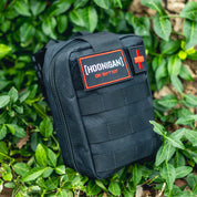Black Hoonigan OH SH*T Safety Kit pouch with red cross patch, placed among green ivy leaves outdoors.