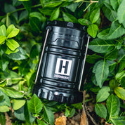A black Hoonigan H.A.V.O.C. lantern with a white logo, resting among green ivy leaves outdoors.