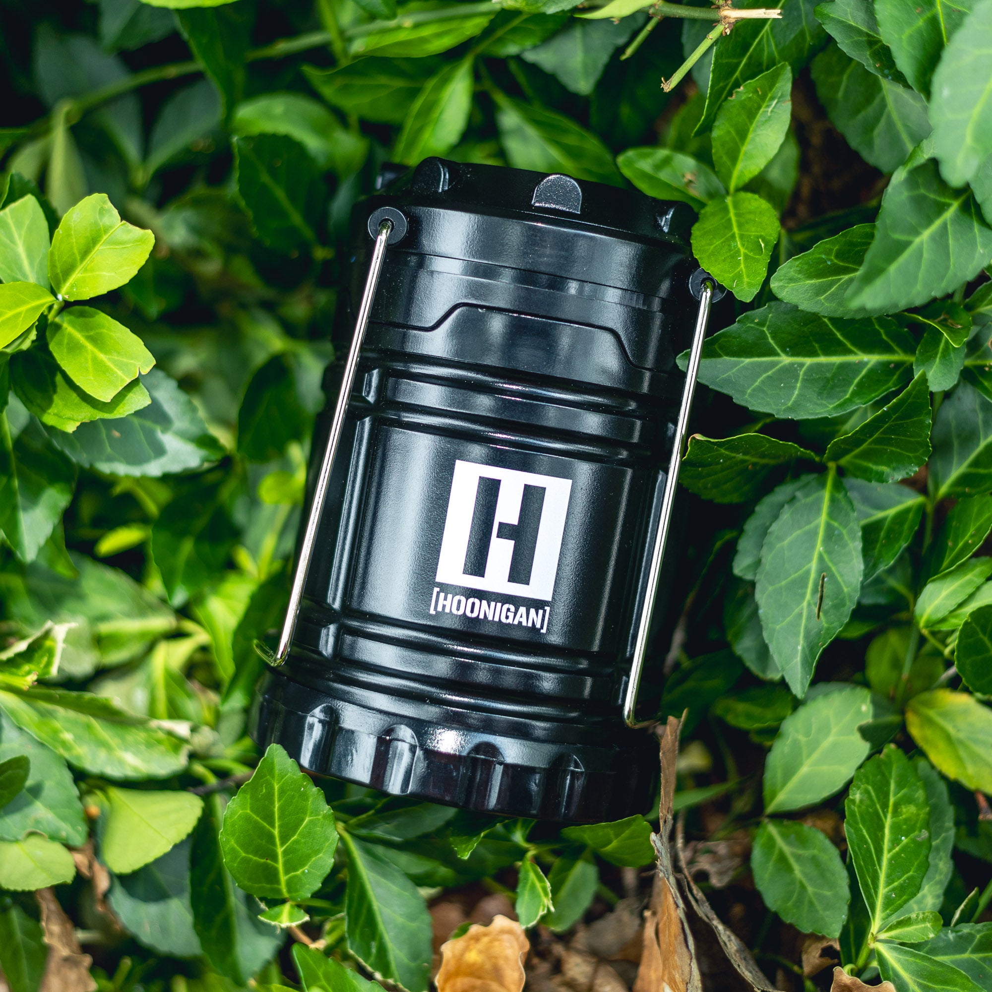 A black Hoonigan H.A.V.O.C. lantern with a white logo, resting among green ivy leaves outdoors.