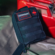 Black Hoonigan OH SH*T Safety Kit pouch with red and white logo, featuring a red cross patch, placed on a surface near a red vehicle.