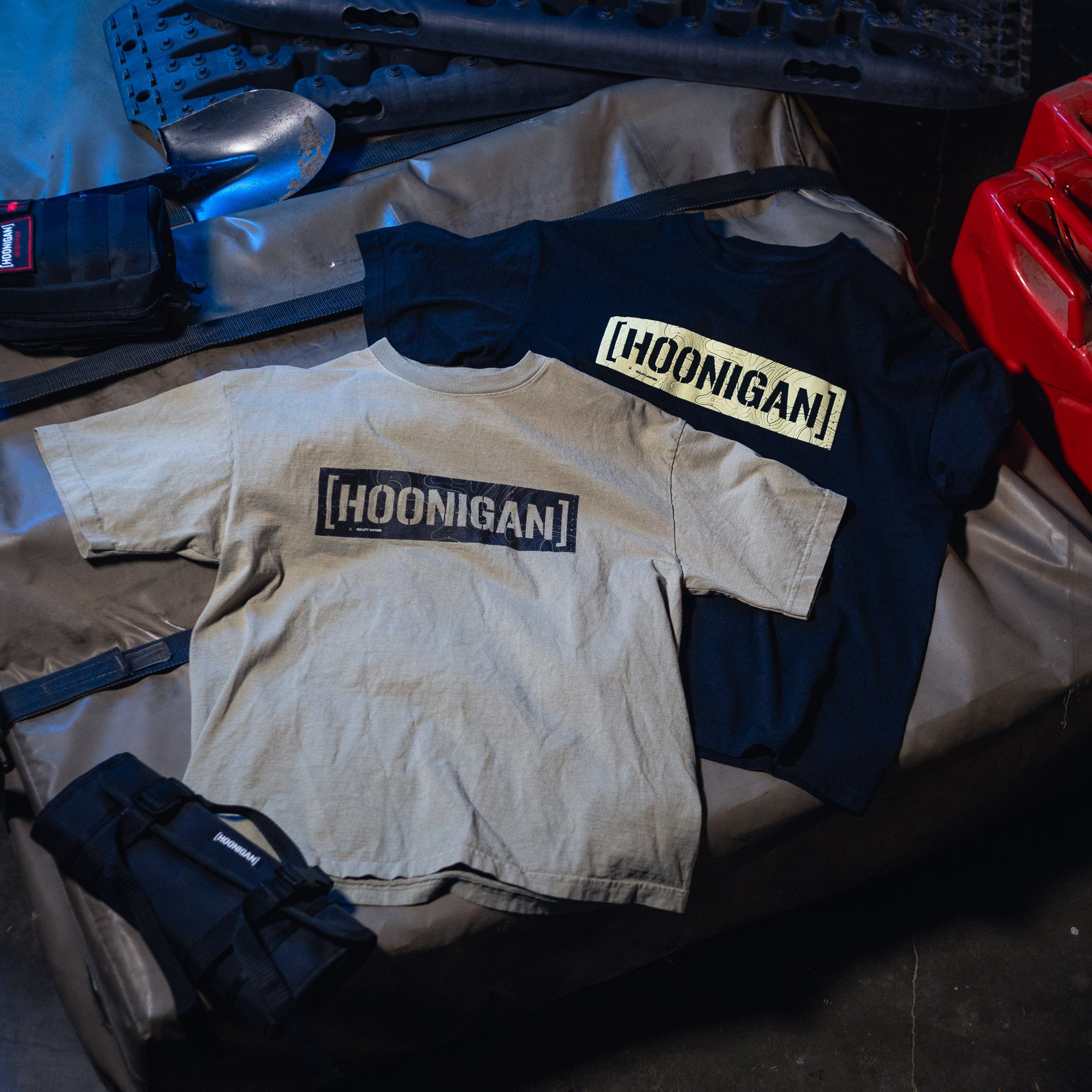 Two Hoonigan TOPOGRAPHIC CENSOR BAR short sleeve tees, one beige and one black, laid flat on a surface with tools and gear nearby.