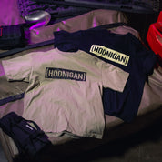 Two T-shirts with "[HOONIGAN]" graphic print, one in beige and one in black, displayed on a surface with gear and equipment.