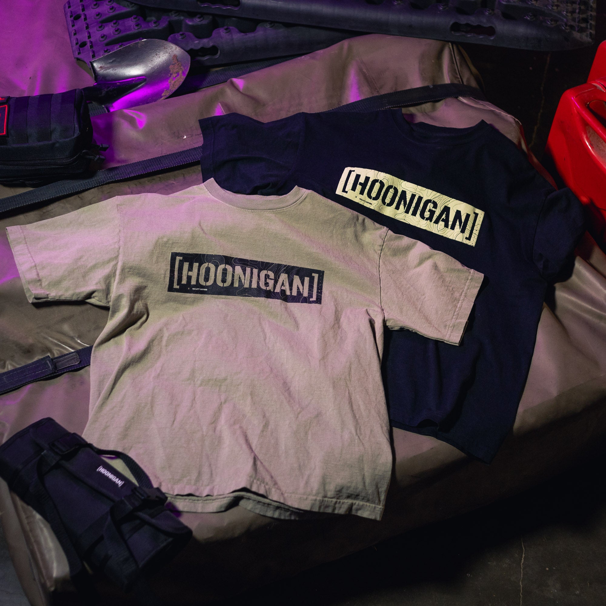 Two T-shirts with "[HOONIGAN]" graphic print, one in beige and one in black, displayed on a surface with gear and equipment.