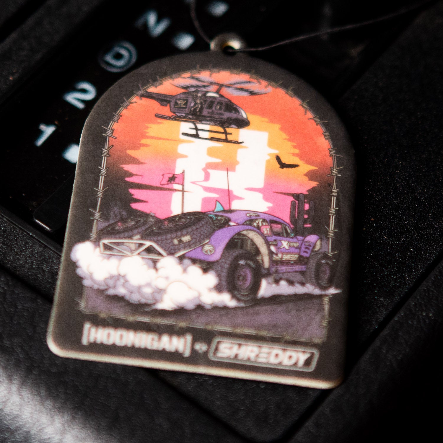 Hoonigan x Shreddy DESERT Air Freshener featuring a vibrant design of a car and helicopter against a colorful sunset backdrop.