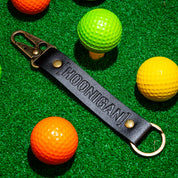 Hoonigan leather keychain featuring a bold embossed logo, set against colorful golf balls on green turf.
