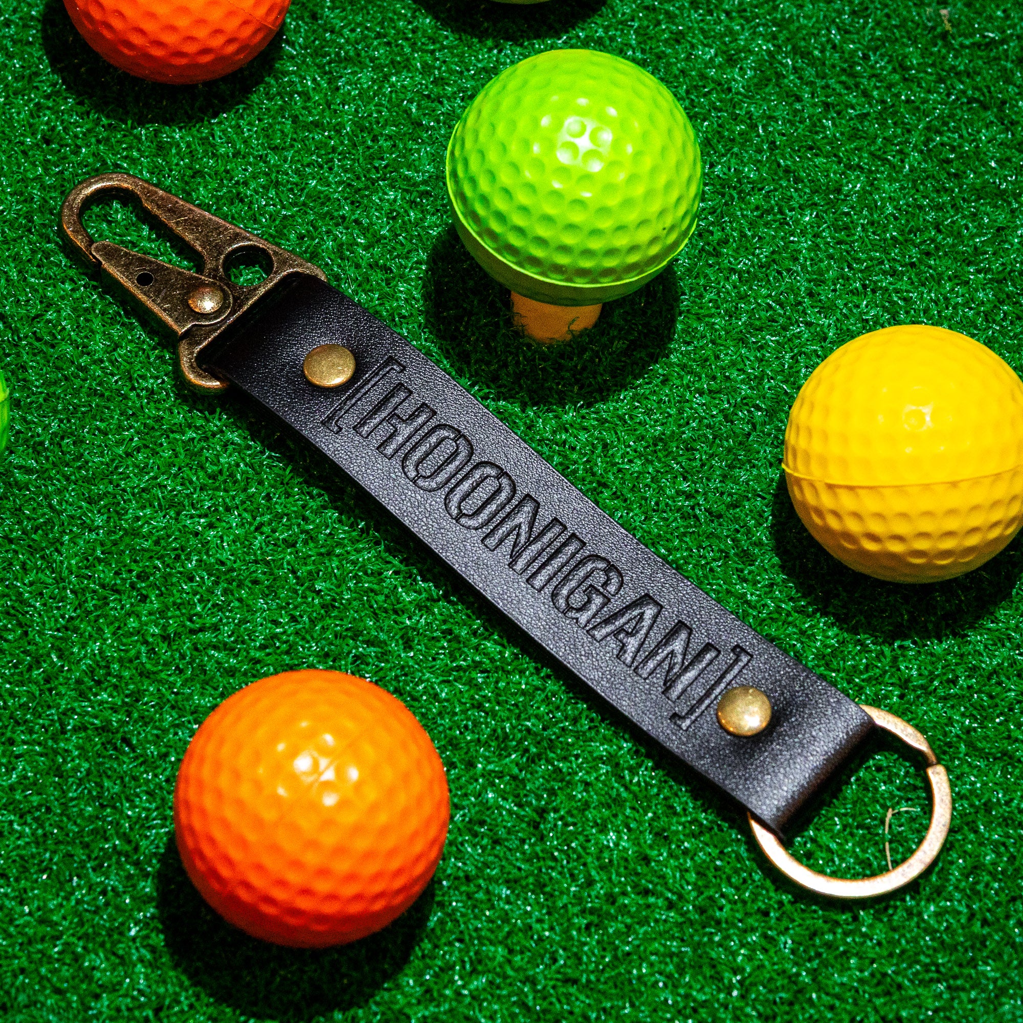 Hoonigan leather keychain featuring a bold embossed logo, set against colorful golf balls on green turf.