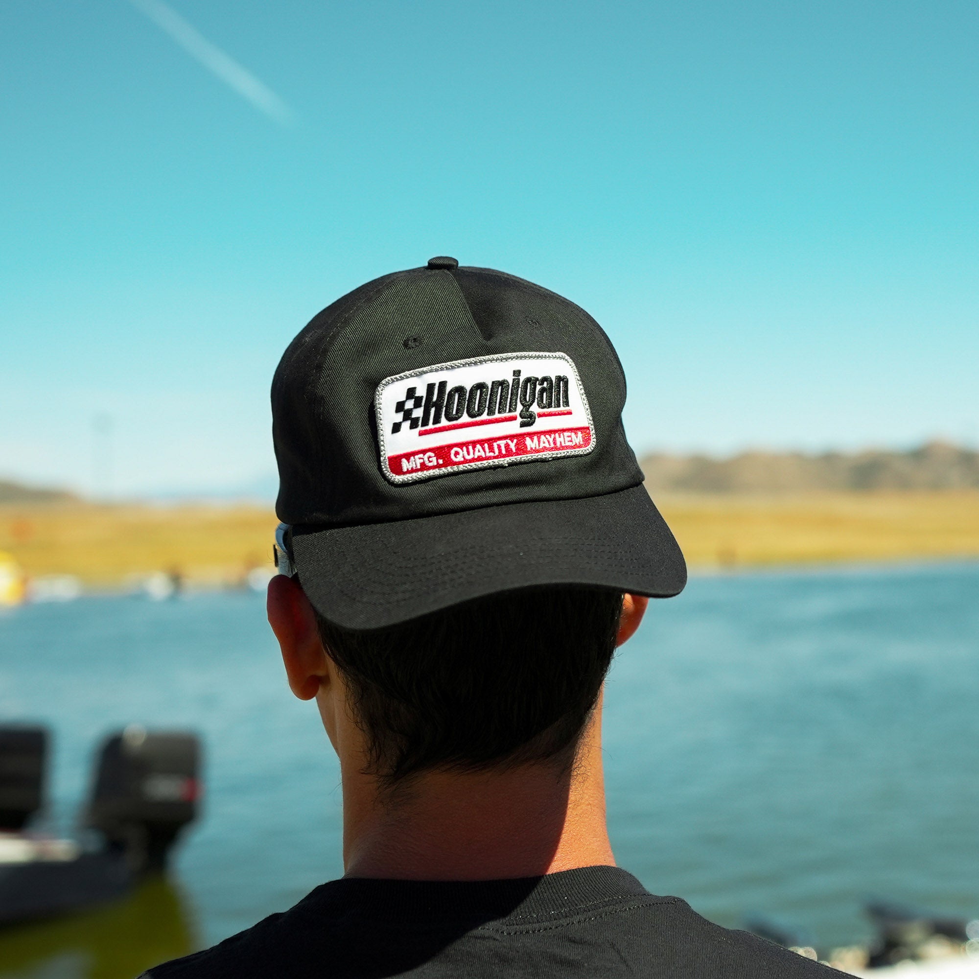 Hoonigan RACEDAY unstructured strapback hat worn by a person facing a lake, showcasing its bold logo and design.
