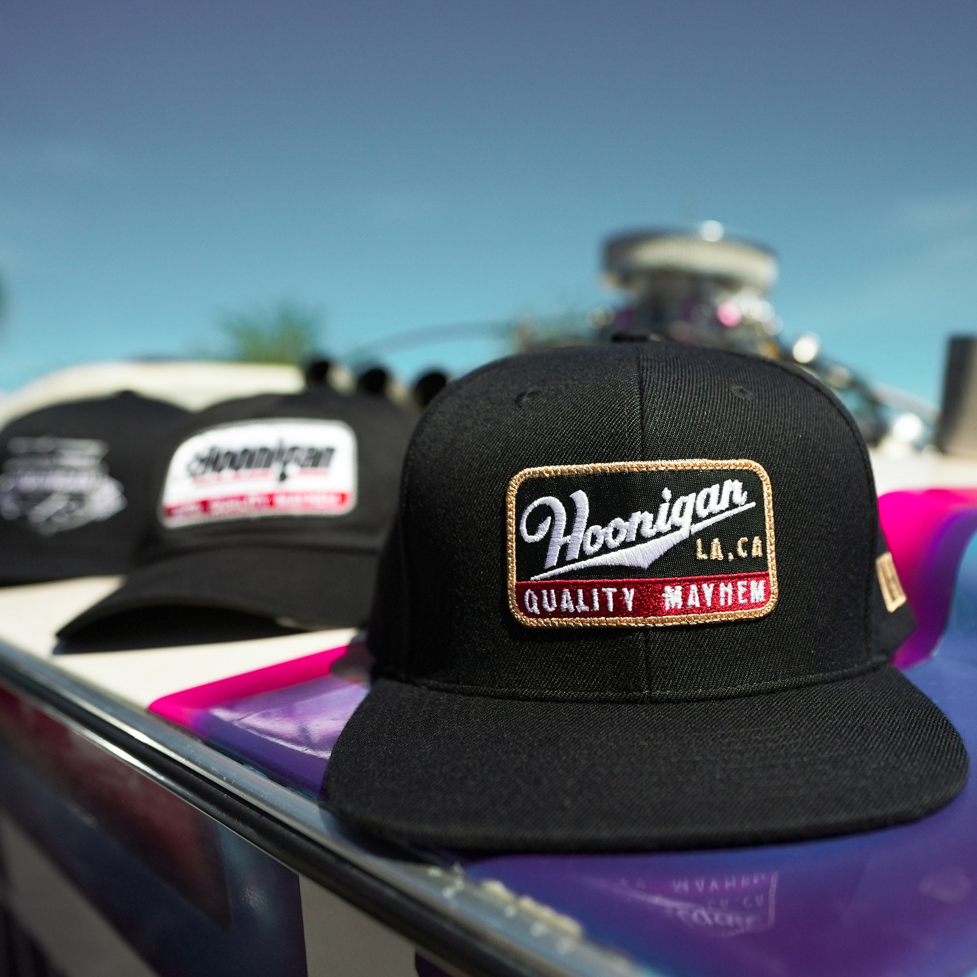 Black Hoonigan Champagne snapback hat featuring embroidered logo, displayed on a colorful surface with other hats in background.