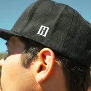 Close-up of a black snapback hat featuring the Hoonigan logo, worn by a person outdoors, showcasing automotive style.
