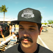 Hoonigan Victory Lap Snapback Hat worn by a man with a mustache, featuring a bold logo against a sunny backdrop.