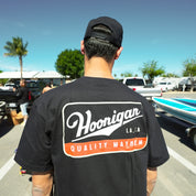 Back view of a man wearing a black Hoonigan CHAMPAGNE short sleeve t-shirt featuring a bold logo and "Quality Mayhem" text.