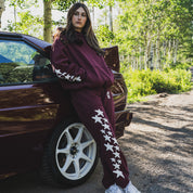 Model wearing Hoonigan x Lia Block UR STAR Pullover Hoodie, posing by a car in a forest setting, showcasing automotive lifestyle.