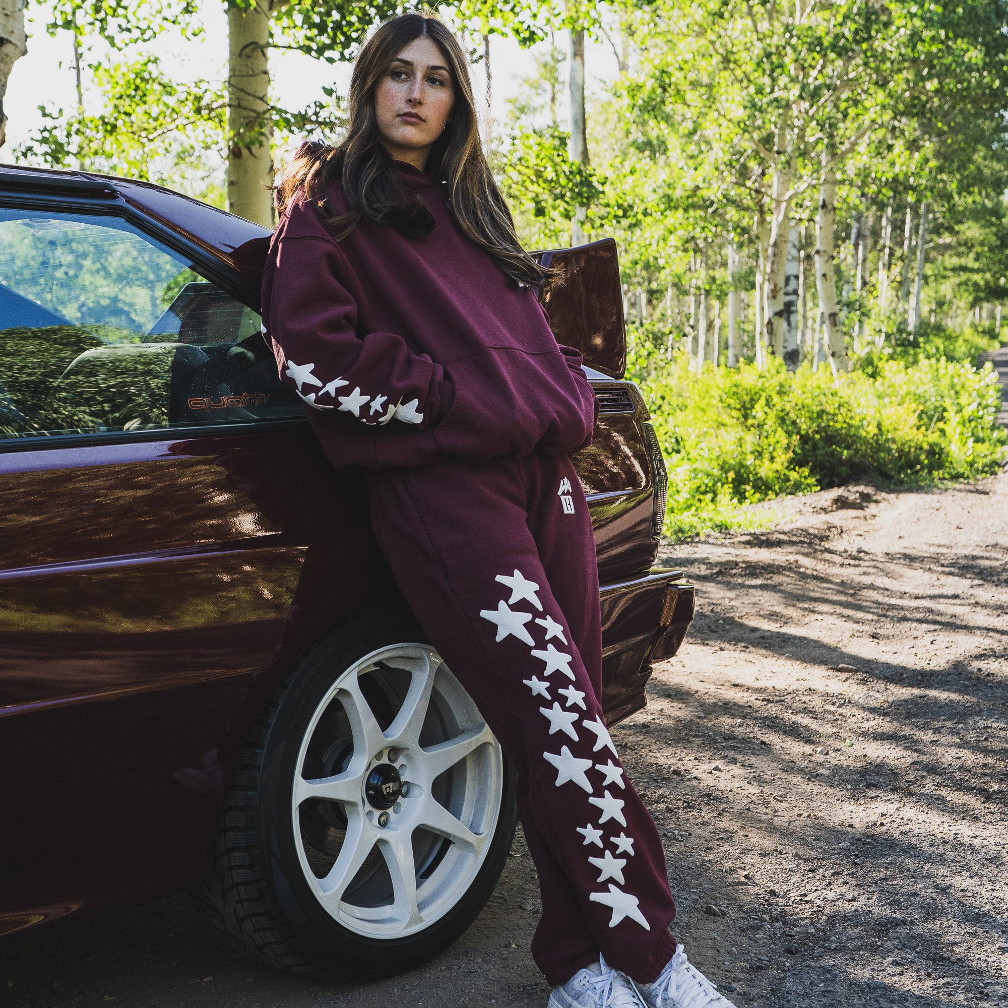 Model wearing Hoonigan x Lia Block UR STAR Pullover Hoodie, posing by a car in a forest setting, showcasing automotive lifestyle.
