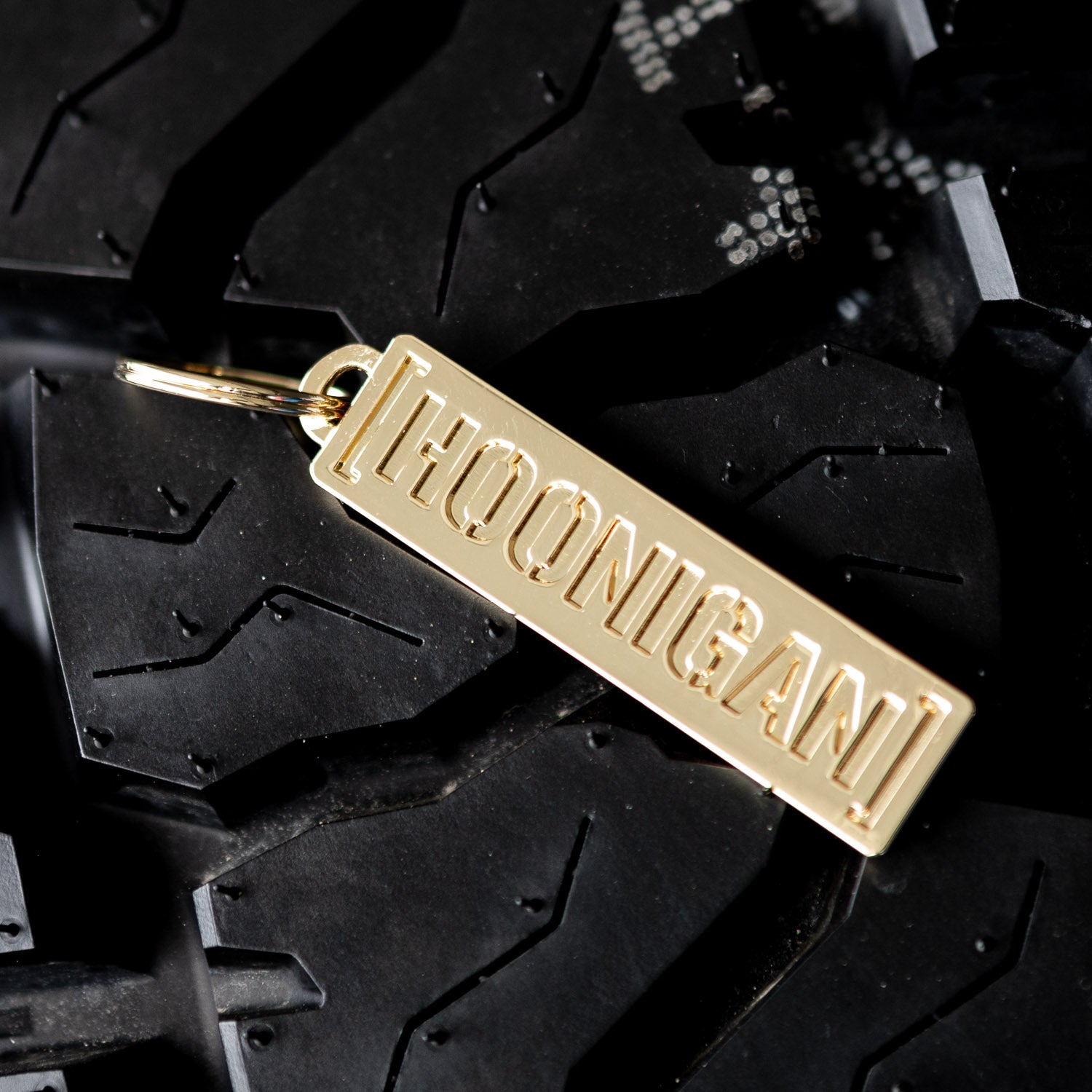 Gold Hoonigan metal censor bar keychain resting on a textured tire tread background, showcasing its sleek design.